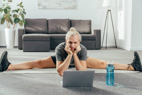 sporty adult man doing split exercise and looking at laptop