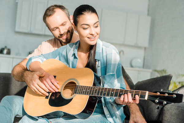happy husband teaching smiling wife to play acoustic guitar