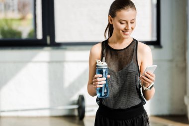 spor şişe su ile tutarak ve spor salonunda smartphone kullanarak sporcumuz gülümseyen