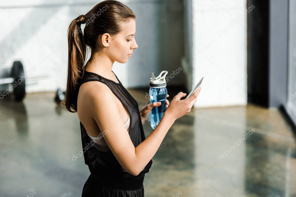 side view of fit sportswoman holding sport bottle with water and using smartphone at gym