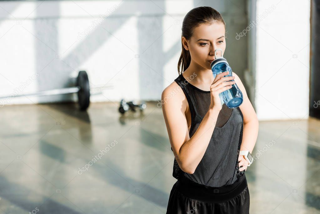 beautiful concentrated sportswoman drinking water from sport bottle at gym