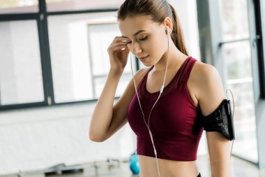 beautiful slim sportswoman in smartphone armband putting on earphones at gym clipart