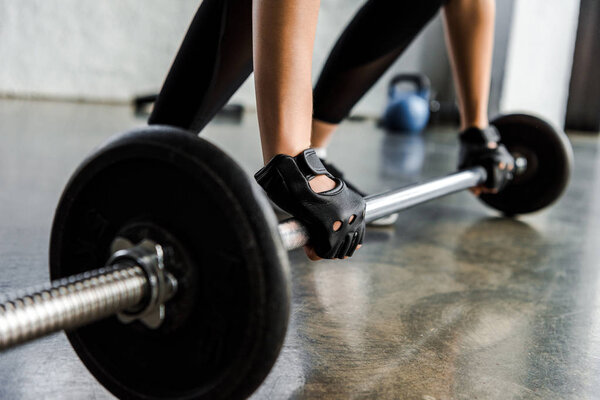 cropped view of sportswoman in weight lifting gloves training with barbell at fitness studio