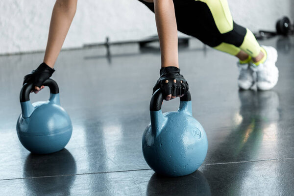 cropped view of sportswoman in weightlifting gloves doing plank exercise on kettlebells at sports center