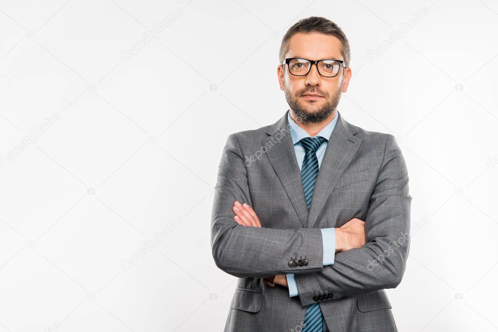 confident businessman in suit and eyeglasses standing with crossed arms and looking at camera isolated on white
