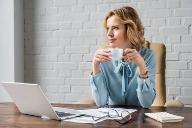 smiling businesswoman holding cup of coffee and looking away while sitting at workplace clipart