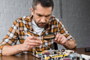 close-up portrait of concentrated electronics engineer with tweezers and magnifying glass repairing motherboard