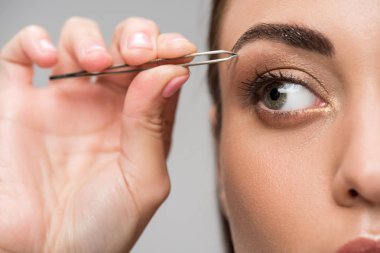close up of young woman correcting shape of eyebrows with tweezers isolated on grey clipart