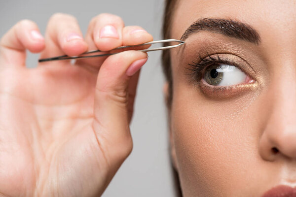 крупным планом молодая женщина корректирует форму бровей пинцетом, изолированным на сером
