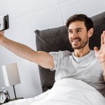 スマート フォンでビデオ チャットを有するベッドで手を振って笑顔の若い男