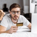 Junger Mann in Brille und Schlafanzug mit Kreditkarte und Laptop auf dem Bett