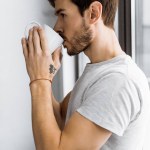 Vista lateral de joven atractivo con taza de café recostado en la ventana en casa