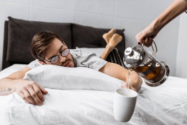 cropped shot of someone pouring tea into cup while sleepy man in eyeglasses lying on bed  clipart