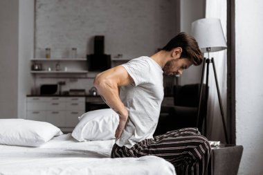 Sabahleyin yatakta otururken sırt ağrısı muzdarip pijama genç adam yan görünüm