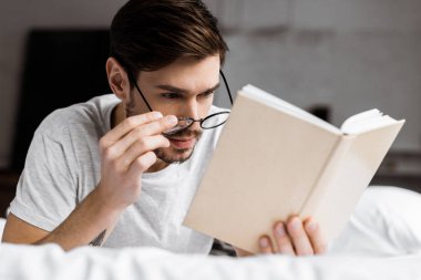 yakışıklı genç adam yatakta yatarken kitap okuma ve gözlük ayarlama