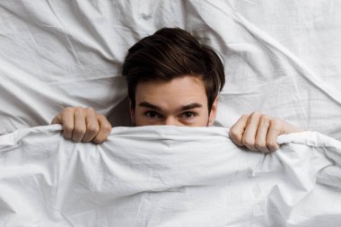 top view of young man hiding under blanket in bed and looking at camera clipart