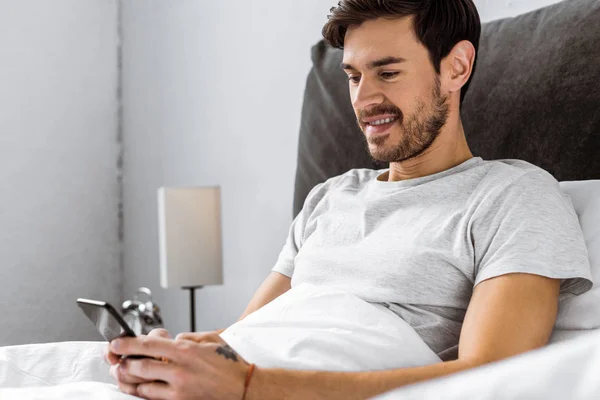 Handsome Smiling Young Man Sitting Bed Using Smartphone — Free Stock Photo