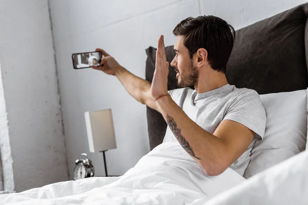 Knappe Jongeman Chatten Met Smartphone Zwaaien Hand Bed — Gratis stockfoto