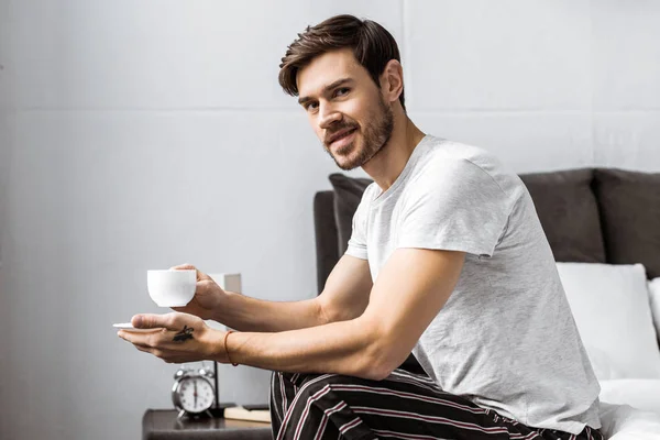 Young Man Pajamas Holding Cup Coffee Smiling Camera While Sitting — Free Stock Photo