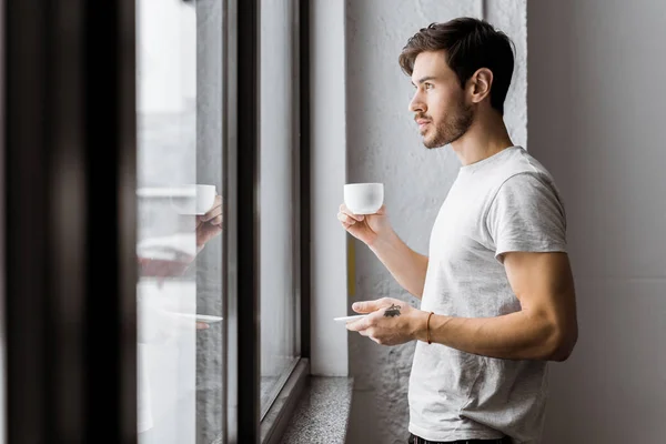 Vista Lateral Del Joven Sosteniendo Una Taza Café Mirando Ventana — Foto de Stock