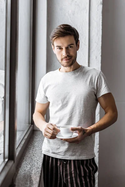 handsome young man in pajamas holding cup of coffee and looking at camera