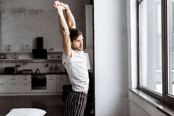 side view of young man in pajamas stretching and looking at window in the morning