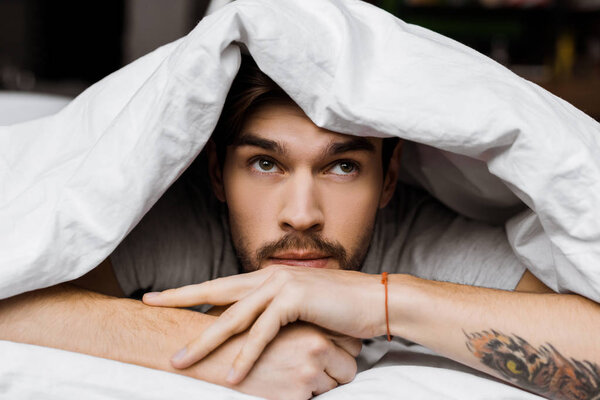 handsome young man lying under blanket in bed at home and looking away