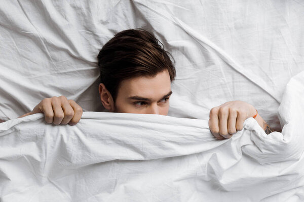 top view of handsome young man hiding under blanket in bed at home and looking away