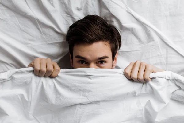 Ovanifrån Ung Man Gömmer Sig Täcket Sängen Och Tittar Kameran — Stockfoto