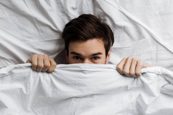 вид сверху на молодого человека, прячущегося под одеялом в постели и смотрящего в камеру
