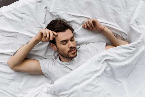 top view of handsome young man lying under white blanket in bed at home