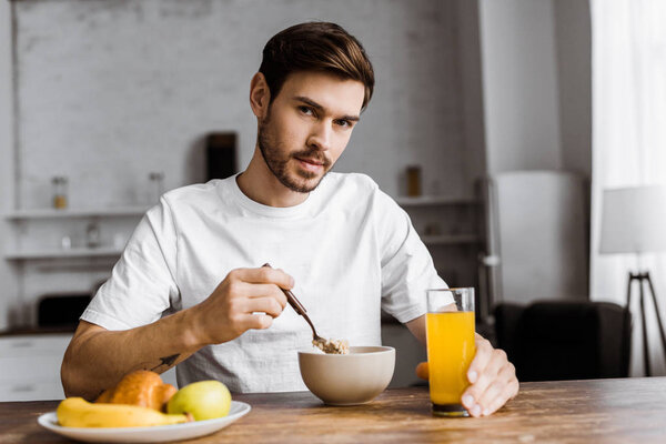 handsome young man having cereal with orange juice and looking at camera at home