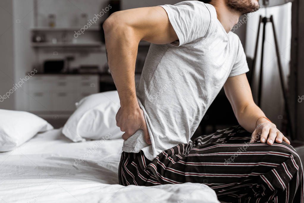 cropped shot of young man in pajamas suffering from backache while sitting on bed in the morning