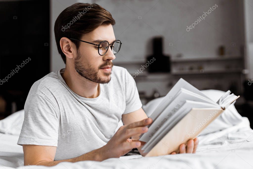 young man in eyeglasses lying in bed and reading book in the morning