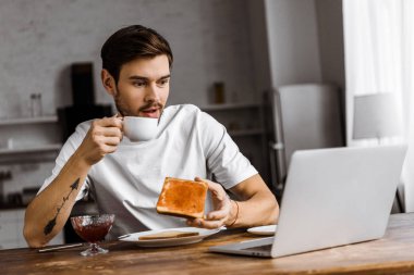 reçel ve kahve ile tost yemek ve evde dizüstü bilgisayar ekranına bakarak şok genç serbest meslek
