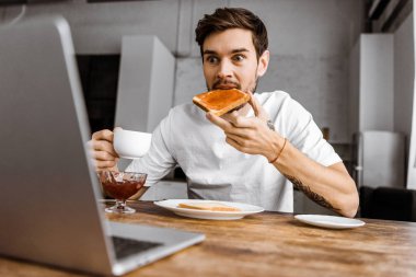 reçelli ekmek yeme ve evde dizüstü bilgisayar ekranına bakarak sürpriz genç serbest meslek