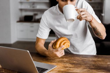 cropped shot of young freelancer with cup of tea, croissant and laptop working on kitchen at home clipart