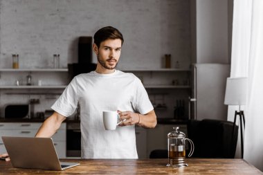 attractive young freelancer with cup of tea and laptop working on kitchen at home clipart