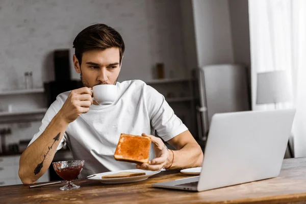 Knappe Jonge Freelancer Eten Toast Met Jam Koffie Kijken Naar — Gratis stockfoto