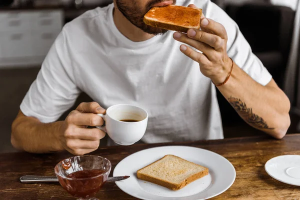 裁剪拍摄的年轻人吃烤面包与果酱和喝咖啡在家里 — 图库照片