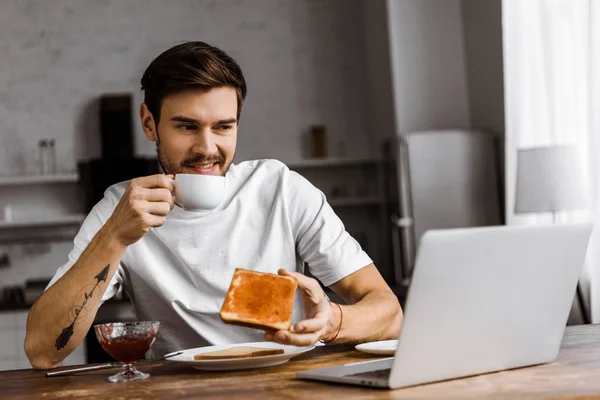 Grimassen Jonge Freelancer Toast Met Jam Eten Kijken Naar Laptop — Gratis stockfoto