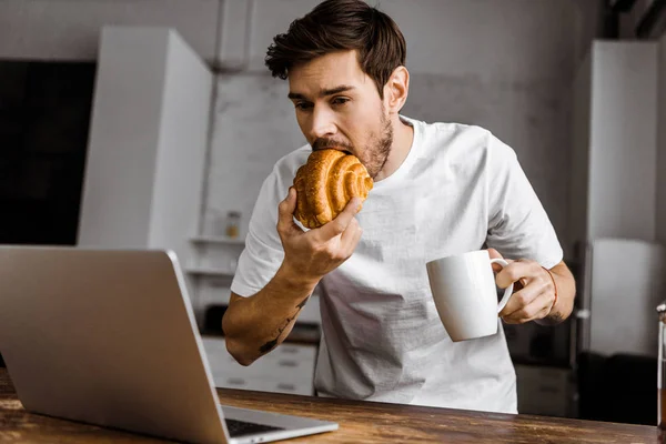 Hungry Young Freelancer Cup Tea Croissant Laptop Working Kitchen Home — Stock Photo, Image