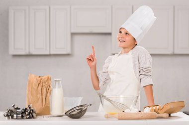 neşeli çocuk chef şapka ve mutfakta masada yemek hazırlama sırasında fikir jest yapıyor önlük