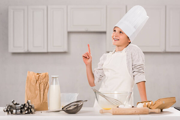 cheerful boy in chef hat and apron doing idea gesture during food preparation at table in kitchen