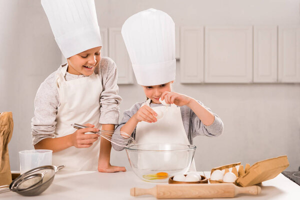 smiling brother and sister in aprons and chef hats during food preparation at table in kitchen 