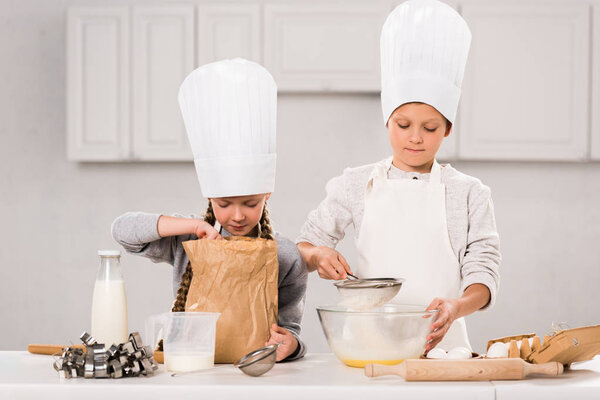 adorable children in aprons and chef hats during food preparation at table in kitchen 