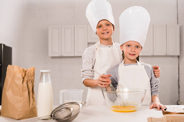 happy sister and brother in aprons and chef hats whisking eggs in bowl at table in kitchen 