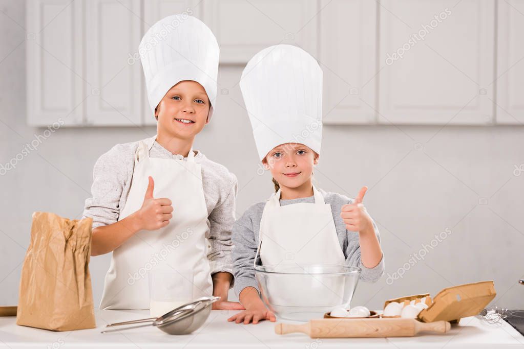 adorable brother and sister in chef hats showing thumbs up during food preparation in kitchen