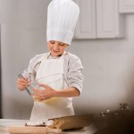 Selective focus of happy boy in chef hat and apron whisking eggs in bowl at table in kitchen