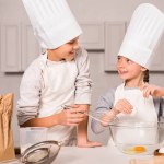 Sœur et frère souriants dans des chapeaux de chef et tabliers fouettant des œufs dans un bol à table dans la cuisine